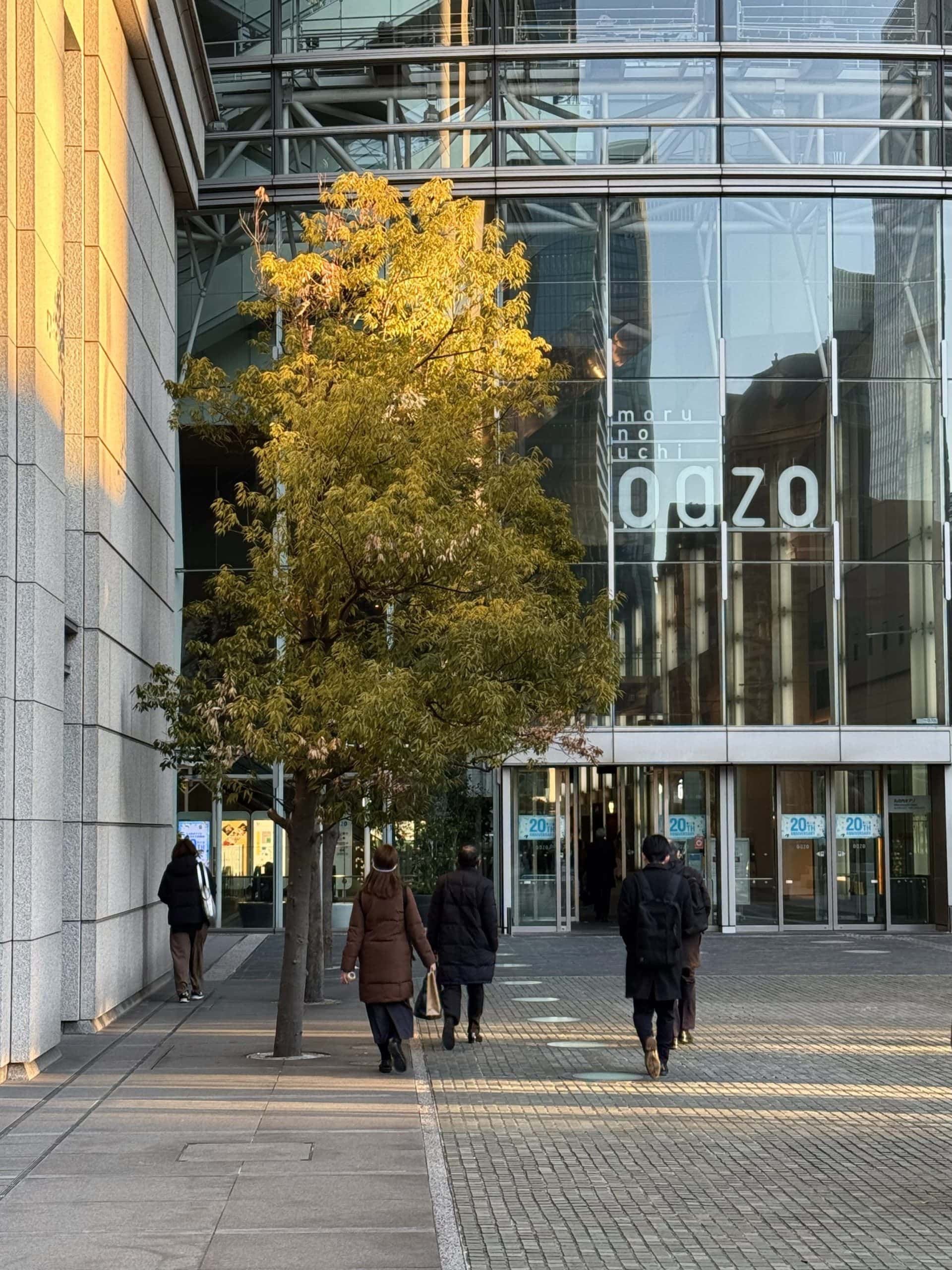 Streaks of morning sunlight hit trees and walls outside the Marunouchi Oazo Building in Tokyo, Japan, as some pedestrians are seen walking towards the building. © 2025 Norbert Woehnl Photography.