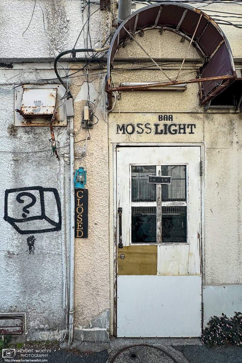 In the Koenji area of Tokyo, Japan, this photo shows a patchwork-looking door that is the entrance to a bar named Moss Light. To the left of the door is a Closed sign and a sprayed black wall graffiti of a black cube with a question mark inside. © 2024 Norbert Woehnl Photography.