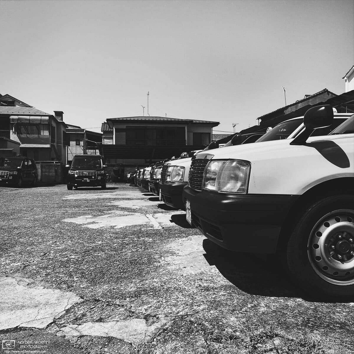 High-contrast monochrome photograph of a line-up of old taxis parked at Tohto Taxi in the Azusawa neighborhood of Itabashi Ward in Tokyo, Japan. © 2022 Norbert Woehnl Photography.