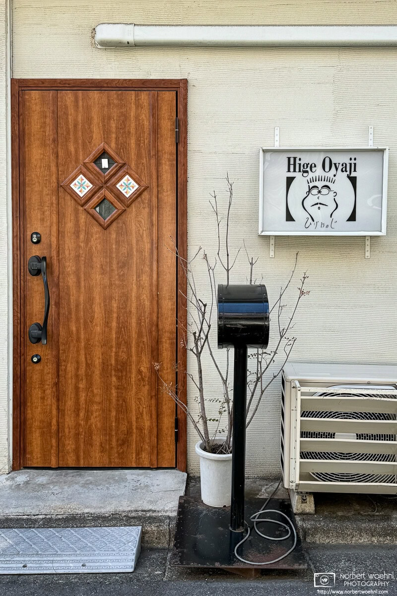 In the Koenji area of Tokyo, Japan, this photo shows an elegant-looking brown residential entrance door. To its right is a shop sign that says Hige Oyaji, which roughly translates to old man with beard. © 2024 Norbert Woehnl Photography.