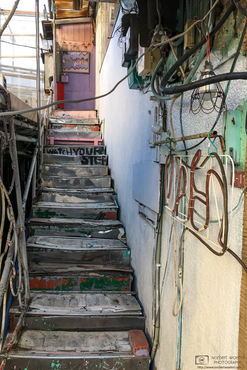In the Koenji area of Tokyo, Japan, this photo shows a very dilapidated and unsafe-looking ascending flock of stairs. The stairs are uneven in shape and size, and on the adjacent wall is an indescribable mixture of wires, cables and pipes. Someone manually painted Watch your step on the upper part of the stairs. © 2019 Norbert Woehnl Photography.