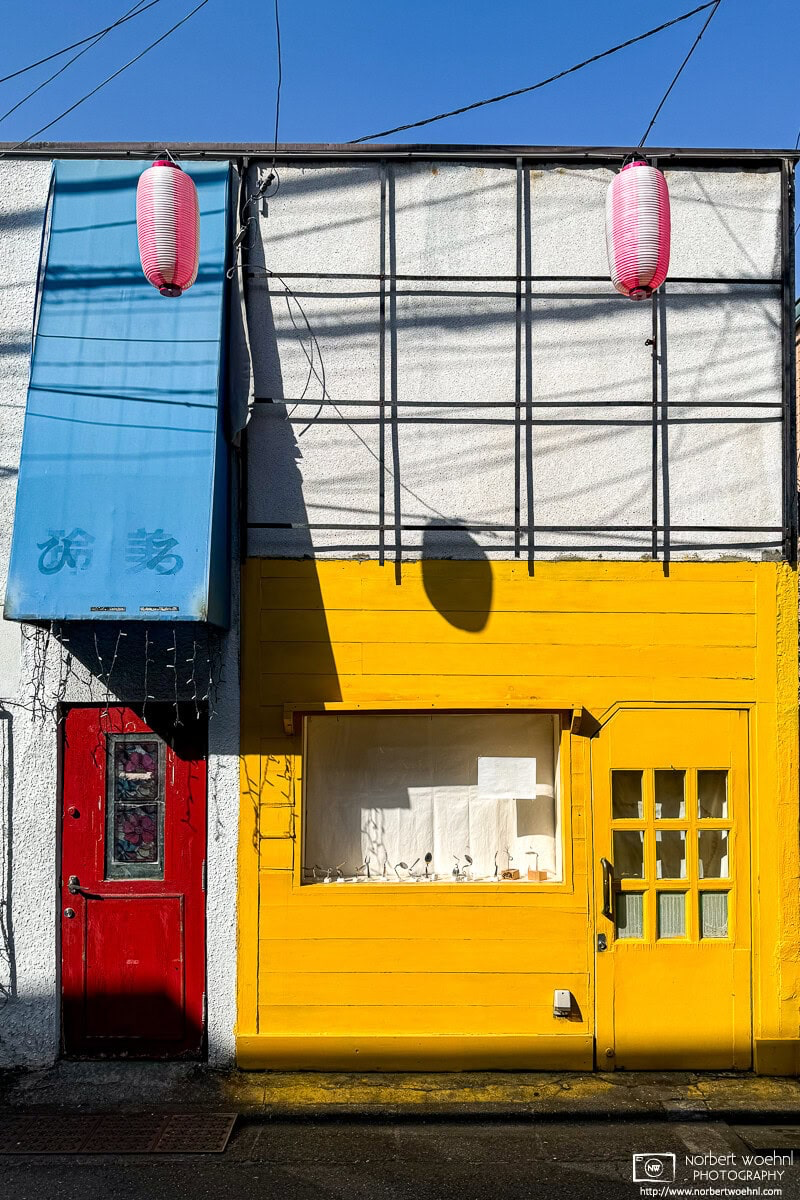 In the Koenji area of Tokyo, Japan, a white-grayish building has two very colorful doors. On the left, a wine-red door covered by a faded blue sun roof, and on the left, a bright yellow door as part of a bigger yellow facade with a window. © 2024 Norbert Woehnl Photography.